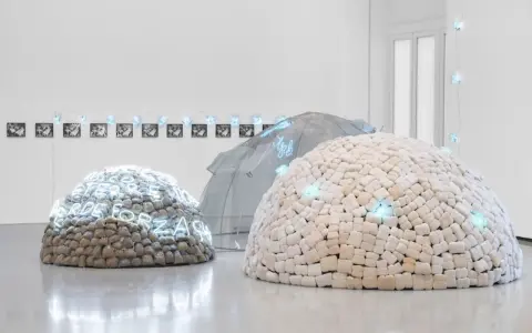 Installation d'art povera : dômes en pierres et en tissu, ornés de néons bleus, sous une structure transparente.
