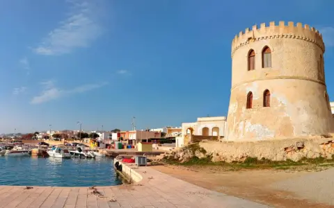 Torre Vado, Lecce: un'antica torre di avvistamento domina un porto con barche e case colorate sotto un cielo azzurro.