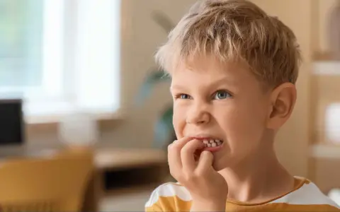Chłopiec z blond włosami i niebieskimi oczami, który gryzie paznokcie. Może to być objaw stresu lub zespołu Tourette'a.
