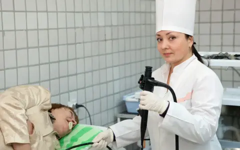 Lekarka w białym fartuchu i czepku przygotowuje pacjenta do gastroskopii, trzymając endoskop.