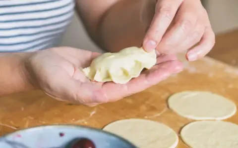 Dłonie formują ciasto na pierogi, tworząc pyszne danie. Na blacie leżą okrągłe placki z ciasta, a obok miseczka z wiśniami.