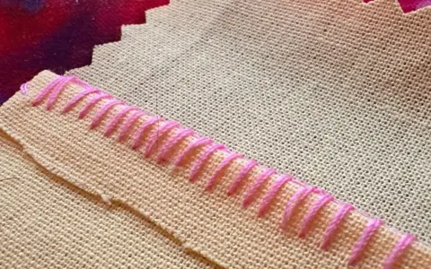 Detalle de tela beige con puntada de hilo rosa formando un borde decorativo.