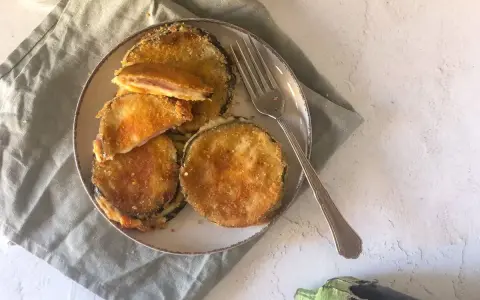 Cordon bleu di melanzane impanati e fritti, ripieni di prosciutto e formaggio filante, serviti su un piatto con una forchetta.