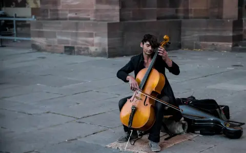Ein Mann spielt auf einem Cello-Musikinstrument vor einer Steinfassade.