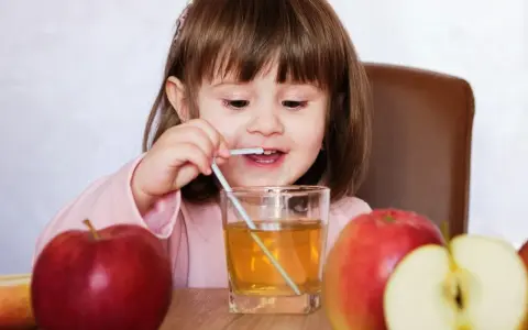 Mała dziewczynka pije sok jabłkowy, który pomaga na zaparcia. Obok stoją jabłka.