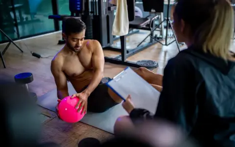 Man med rosa boll på gymmatta, tränare med clipboard. De följer sitt gymprogram.