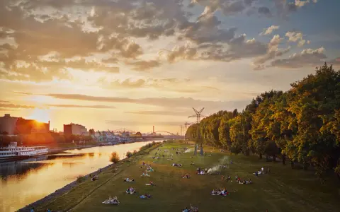 Sonnenuntergang an der Neckarwiese Mannheim. Menschen entspannen im Gras, während die Sonne hinter Industriegebäuden untergeht.
