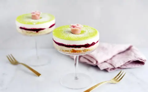 Två vackra prinsesstårtor i glas, med lager av sockerkaka, sylt och grädde, toppade med grön marsipan och en marsipanros.