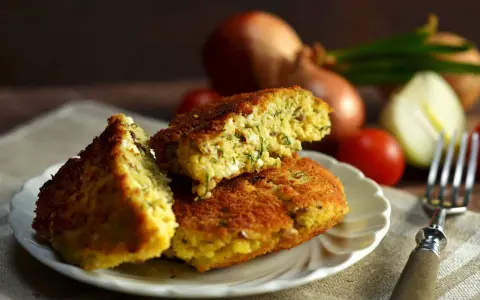 Pyszny kotlet jajeczny z ziołami, podany na talerzyku. W tle cebula i pomidorki.