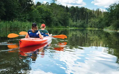 Dwie osoby płyną pomarańczowym kajakiem po spokojnej rzece. Wokół bujna zieleń drzew i trzcin. Idealny spływ kajakowy Mazury.