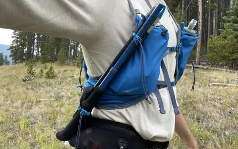 Jak przypiąć kijki do plecaka? Widok z boku na turystę w lesie, z kijami trekkingowymi przymocowanymi do niebieskiego plecaka.