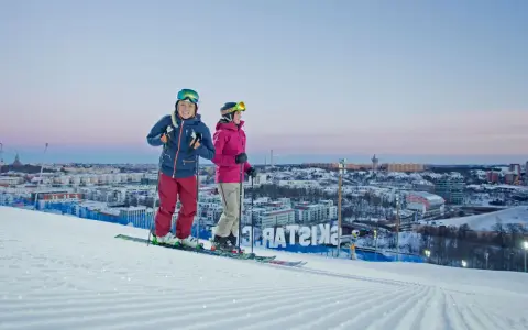 Två skidåkare njuter av utsikten över Hammarbybacken och staden i skymningen.