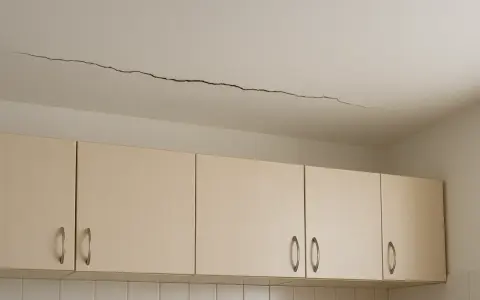 Długie pęknięcie na suficie nad jasnymi szafkami kuchennymi. Wygląda, jakby krzywe ściany zaczęły się odsuwać.