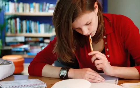 Dziewczyna w czerwonym swetrze myśli nad tekstem rozprawki maturalnej, trzymając długopis przy ustach.