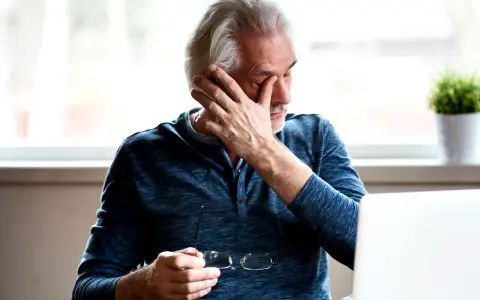Uomo anziano con mano sull'occhio, preoccupato per i vitrectomia rischi. Tiene occhiali, laptop e pianta sullo sfondo.