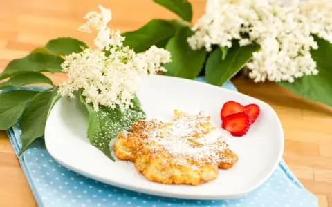 Fiori di sambuco fritti, spolverati di zucchero a velo, serviti su un piatto bianco con fragole fresche e decorati con fiori di sambuco.
