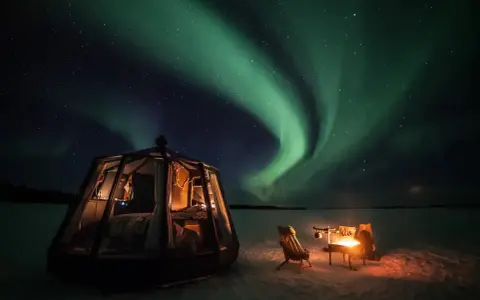 En glaskupol för camping i Norrland lyser upp natten under ett dansande norrsken. En eld sprakar vid sidan om.