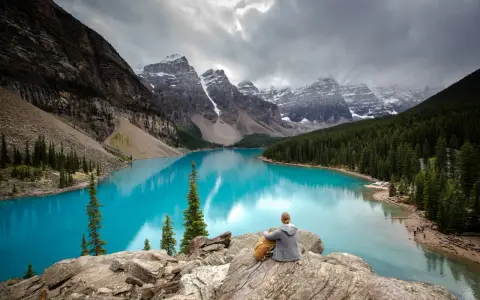 Person sitzt auf Felsen und blickt auf den türkisfarbenen Moränen-See im Banff National Park, Kanada. Majestätische Berge und Nadelwälder umgeben das Wasser.