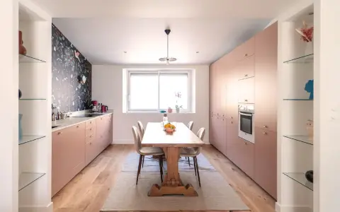 Une cuisine salle à manger et salon dans la même pièce, avec des meubles roses, un papier peint floral et une table en bois.