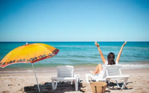 Kobieta cieszy się urlopem wypoczynkowym na plaży, z rękami w górze. Obok leżaki i parasol.