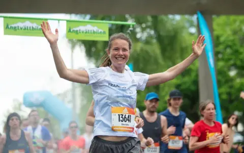 Glad kvinna med armarna uppe vid målgången i ett maraton. En lycklig löpare som njuter av sin maraton tid.