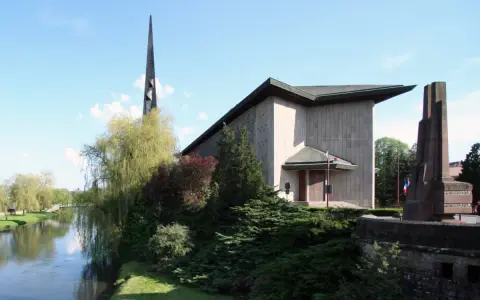 L'église Saint-Rémy de Baccarat, avec sa flèche élancée, se dresse près d'une rivière bordée d'arbres verdoyants.