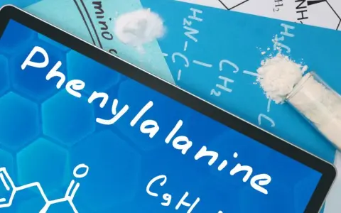 Na ekranie tabletu nazwa "Phenylalanine" i wzór chemiczny. Obok proszek fenyloalaniny w probówce i na kartce.