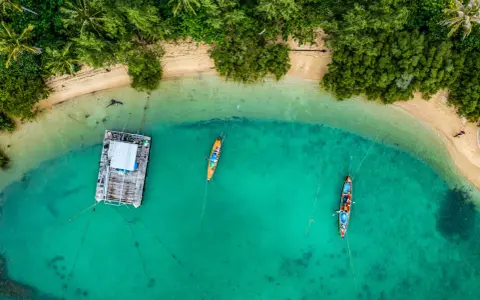 Drohne über Thailand: Ein Blick von oben auf eine schwimmende Plattform und zwei Boote im türkisfarbenen Meer, umgeben von üppiger Vegetation und Sandstrand.