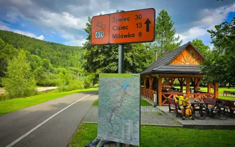 Znak szlaku rowerowego 611 Velo Soła wskazuje kierunki. Obok mapa i wiata z rowerzystą.