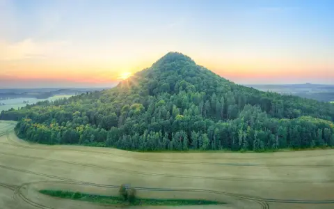 Wygasłe wulkany Dolny Śląsk o poranku. Słońce wschodzi nad zalesionym stożkiem, otoczonym polami.