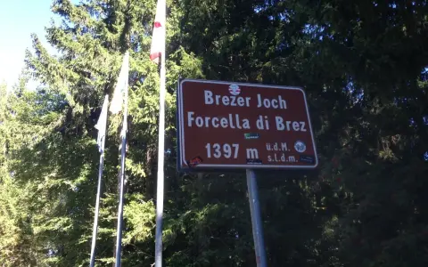Cartello indicante "Forcella di Brez" a 1397 m, con bandiere e alberi sullo sfondo.