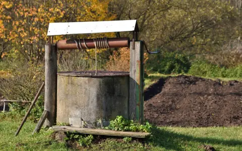 Stara studnia kręgowa z drewnianą konstrukcją i metalowym kołowrotem, otoczona jesienną zielenią i stertą ziemi.