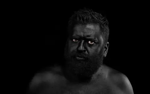 Hombre con el rostro y cuerpo cubiertos de pintura negra, ojos penetrantes. Parece un guerrero ancestral, don fluor.