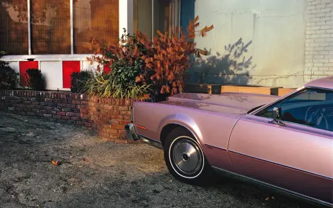 Un'auto rosa pastello, un classico stile William Eggleston, parcheggiata vicino a un muro di mattoni e un edificio con pannelli rossi e bianchi.