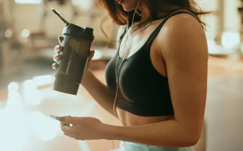 Kobieta w sportowym topie trzyma shaker, zastanawiając się, kiedy pić białko przy odchudzaniu. Słucha muzyki na telefonie.