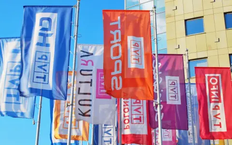 Kolorowe flagi z napisem "TVP w likwidacji" powiewają przed nowoczesnym budynkiem.