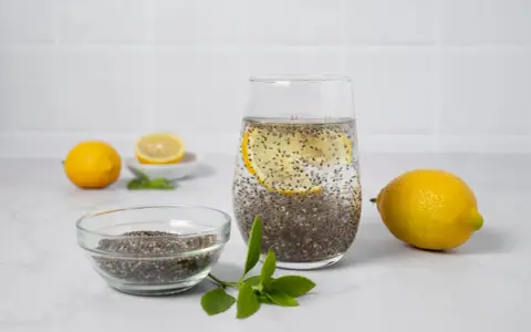 Vaso con agua de chia, rodaja de limón y semillas de chía en un bol. Limones y hojas verdes completan la escena.
