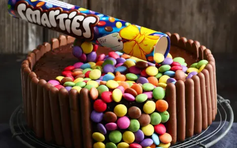 Eine einfache Geburtstagstorte für Jungs: Schokoladenkuchen, umhüllt von Schokoriegeln und bedeckt mit bunten Smarties, die aus einer aufgeschnittenen Rolle rieseln.