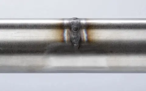 Dettaglio di una saldatura acciaio inox su un tubo metallico, con sfumature iridescenti intorno al cordone.