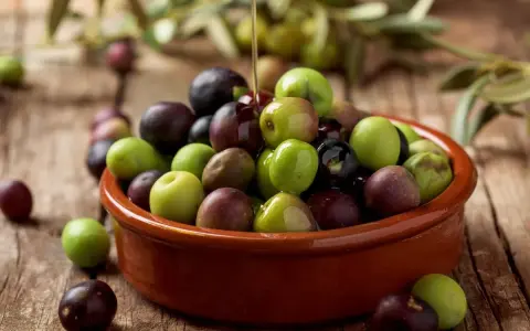Aceite de arbequina vertiéndose sobre aceitunas verdes y moradas en un cuenco de barro. Ideal para ensaladas y aliños.