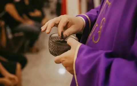A priest in a purple robe takes ashes from a small bowl, preparing for the first day of Lent prayer.