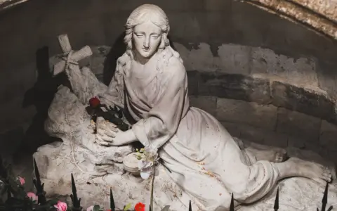 A stone sculpture depicts the serene body of Mary Magdalene, resting with a cross and a single red rose.