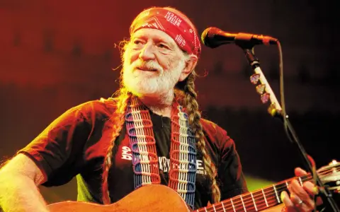 Willie Nelson mit roter Bandana und Zöpfen spielt Gitarre. Ein Mikrofon steht bereit für den Titel von Willie Nelson.