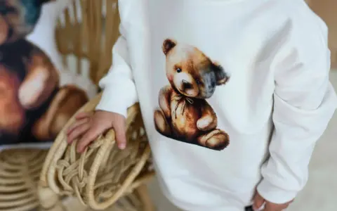 Biała bluza Moschino z uroczym misiem w stylu vintage. Dziecko trzyma się wiklinowego koszyka.