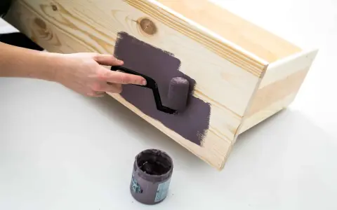 En hand använder en liten roller för att måla en trälåda med mörklila färg, vilket ger en jämn yta utan penseldrag.