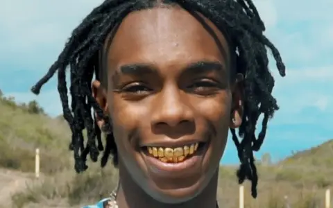 YNW Melly, uśmiechnięty z złotymi zębami, na tle nieba i wzgórz.