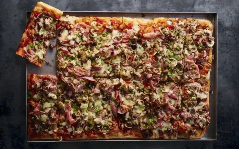 Prostokątna pizza 4 sery z pieczarkami, szynką i oliwkami, pokrojona na kwadraty, z jednym kawałkiem odłożonym na bok.