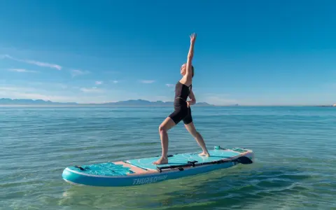 Kvinna utövar sup yoga på en paddleboard i lugnt vatten med berg i bakgrunden.