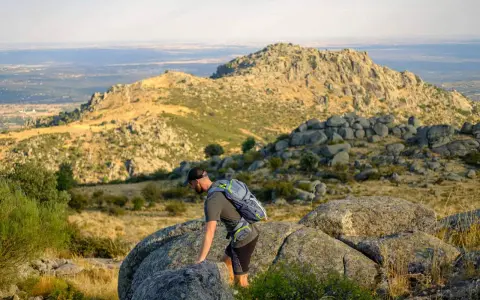 Un excursionista con mochila asciende por un terreno rocoso en la ruta Las Machotas, con un paisaje montañoso y un valle al fondo.