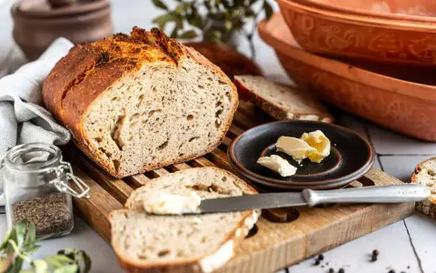Ein rustikales Roggenbrot mit Sauerteig, aufgeschnitten und mit Butter bestrichen. Daneben ein Glas Kümmel und ein Messer.